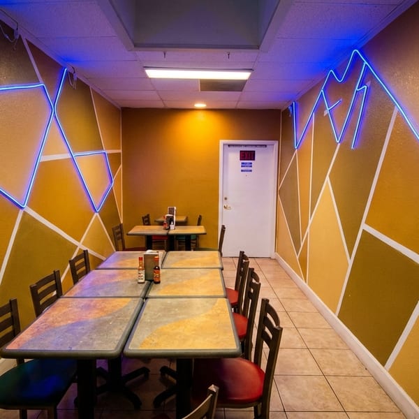 Restaurant dining area with geometric yellow walls, blue neon lights, and multiple tables with chairs.