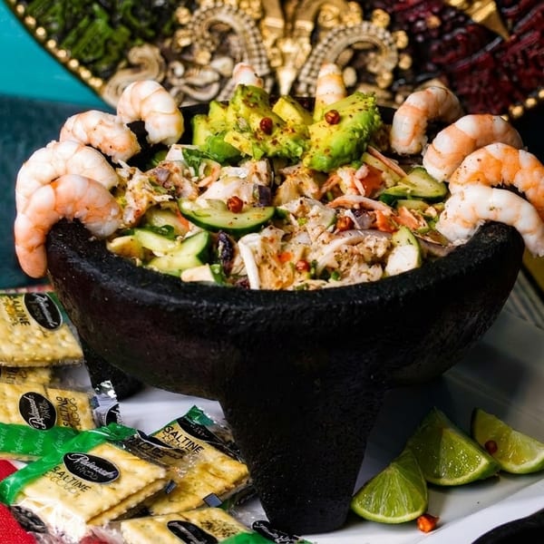 Shrimp and avocado ceviche in a black molcajete, surrounded by crackers, cheese, and lime wedges.